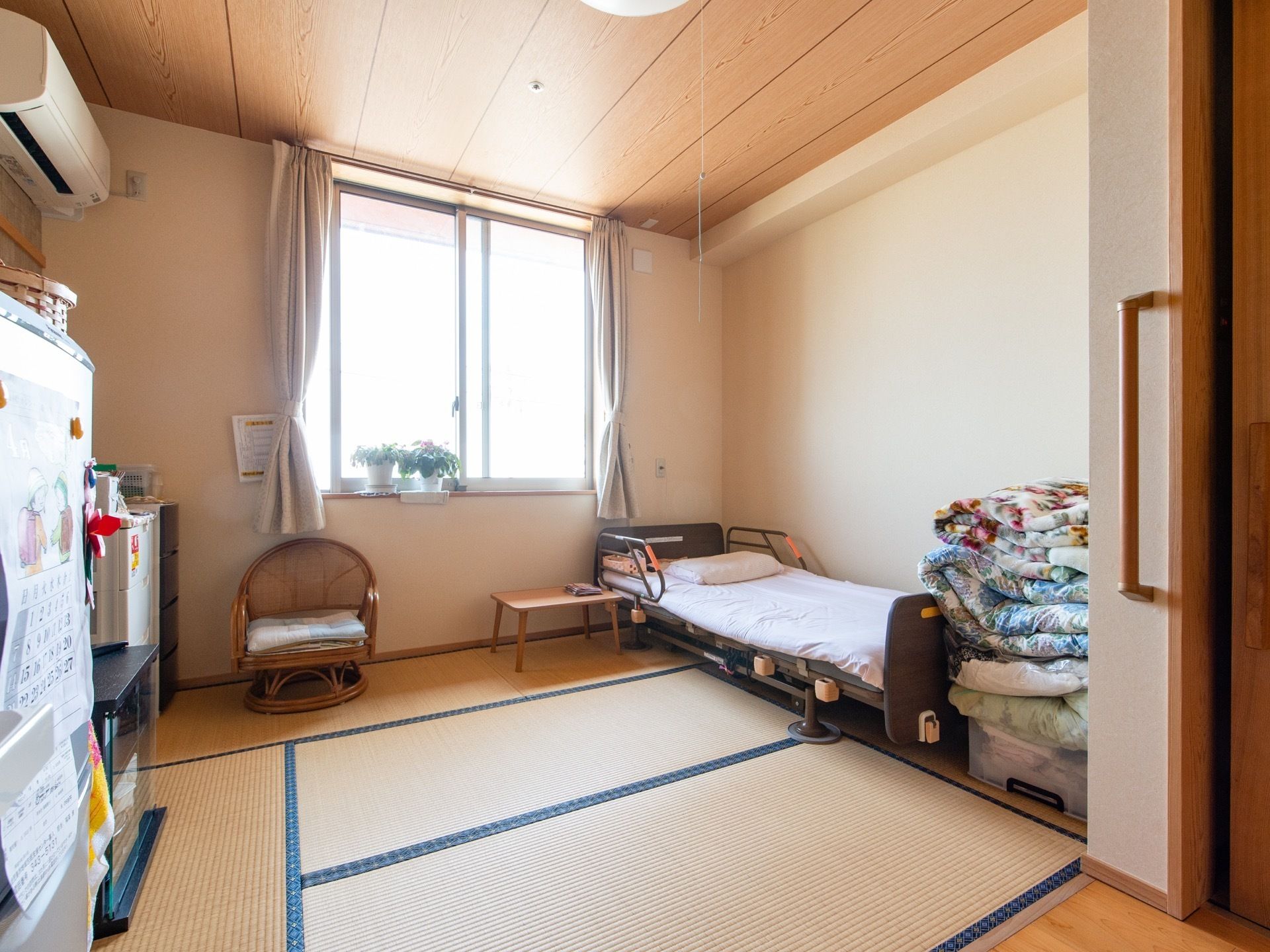 居室のリビングは畳敷きになっている。ベッドやエアコン、冷蔵庫などが設置されている。窓からの採光で明るい。