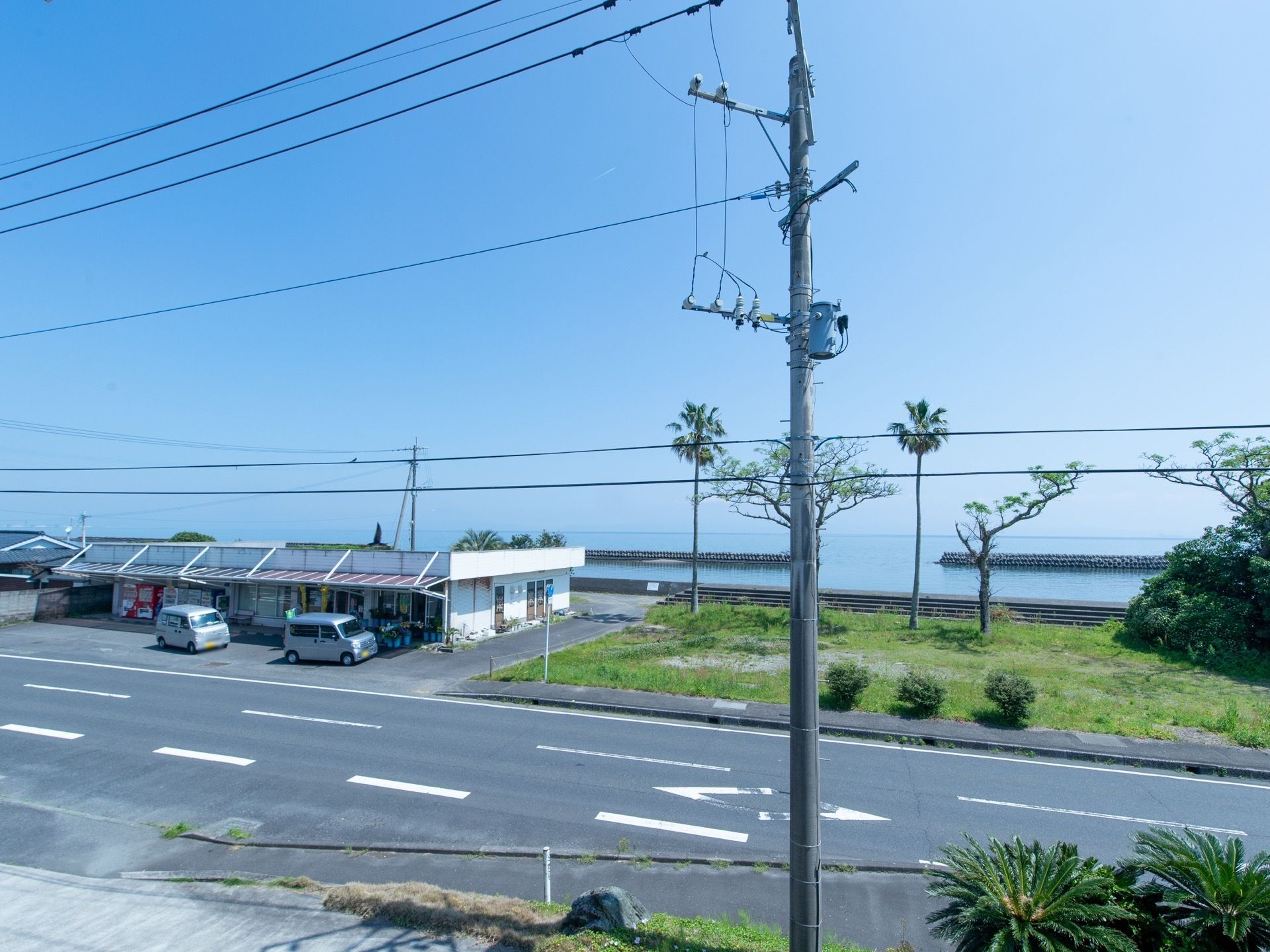 施設からは、道路を隔てて海が見える。道路沿いには、自動販売機を備えた商店があり、車が２台停まっている。