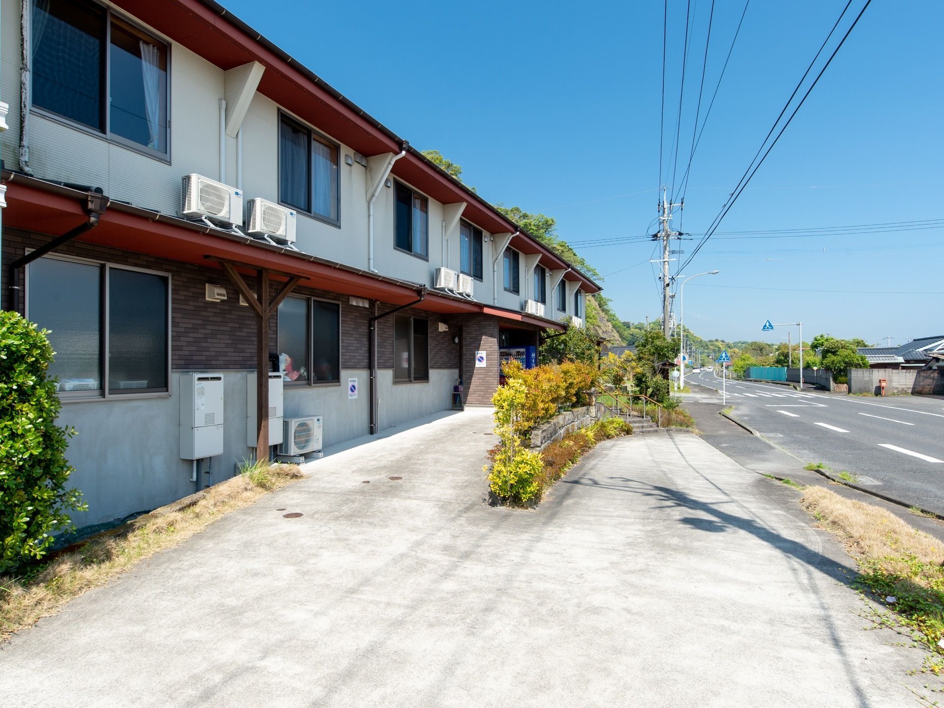 道路に面した立地の２階建ての施設である。建物のすぐそばには、背の高い木が何本か見える。ひさし部分にエアコンの室外機が設置されている。