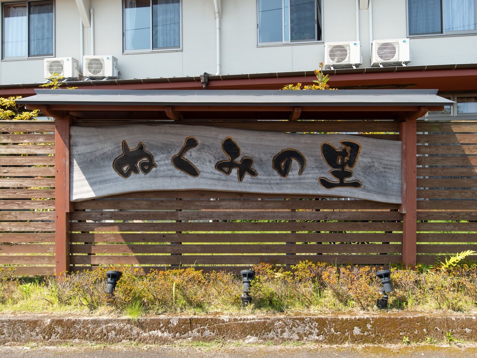 ウッドフェンスの前に、小屋根のついた看板が設置されている。横長の一枚板に大きな文字で施設名称が表示されている。