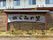 ウッドフェンスの前に、小屋根のついた看板が設置されている。横長の一枚板に大きな文字で施設名称が表示されている。