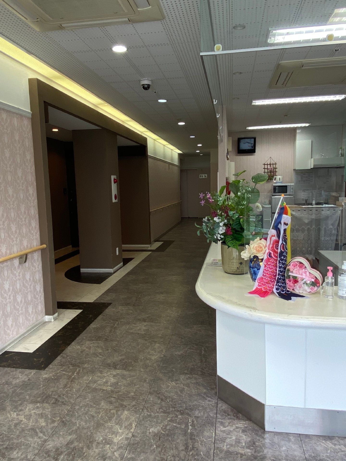 黄色いカーディガンを着た高齢女性が車いすに座り、誰かに向かって手を振っている。そばには女性スタッフがいる。