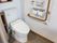 トイレの床面に灰色のカーペットが敷かれている。壁に設置されたＬ字型の手すりのそばに、呼び出しボタンがある。