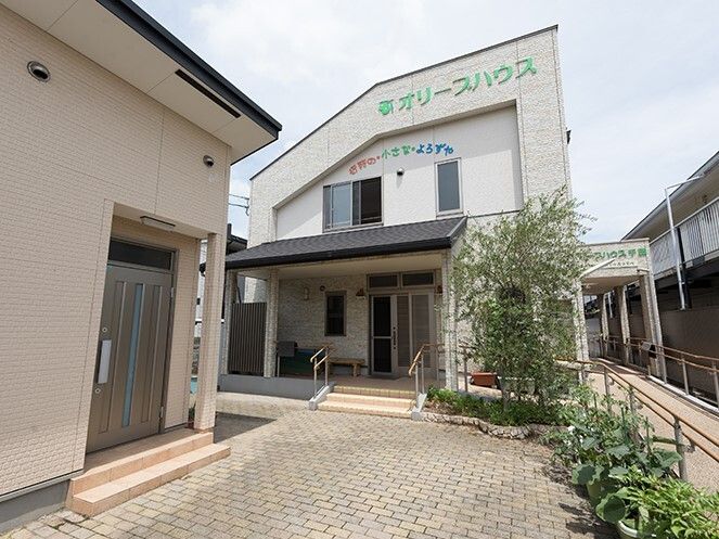 菜園スペースがある2階建ての建物。白を基調としていて、外壁には住宅名が書かれ、玄関からは手すり付きの階段がある。