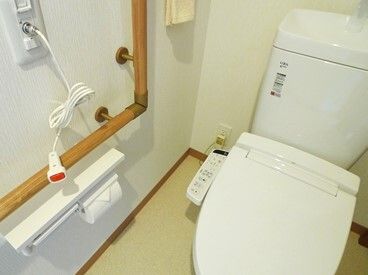 タオルハンガーやエル字型の手すり、呼び出しボタンが設置されたトイレ。便座は温水洗浄ができるタイプで、操作ボタンが便座に付いている。