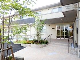 2階建ての建物は緑に囲まれ、玄関には手すり付きのスロープや階段があり、グレーと白で落ち着いた雰囲気。