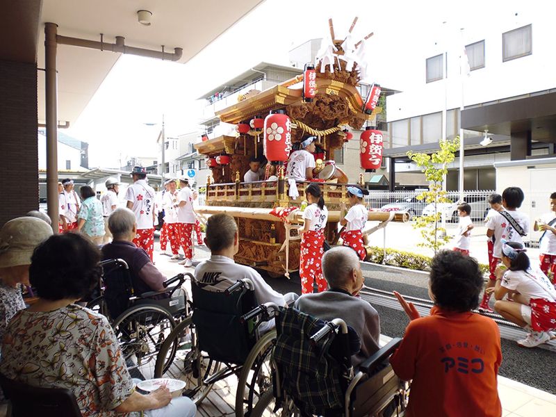 提灯がぶら下がった大きな神輿が玄関前に立ち寄っていて、子どもたちが休憩をしている。そばには車いすの入居者がいる。