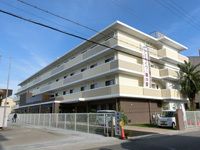 緑が周囲に植えられたベージュ系の外壁の４階建ての建物。玄関前には駐車場も設置されている。