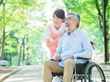 眼鏡をかけた車いすの高齢男性にピンクのエプロンを付けた女性スタッフが付き添い、緑の多い屋外で笑顔で過ごしている。