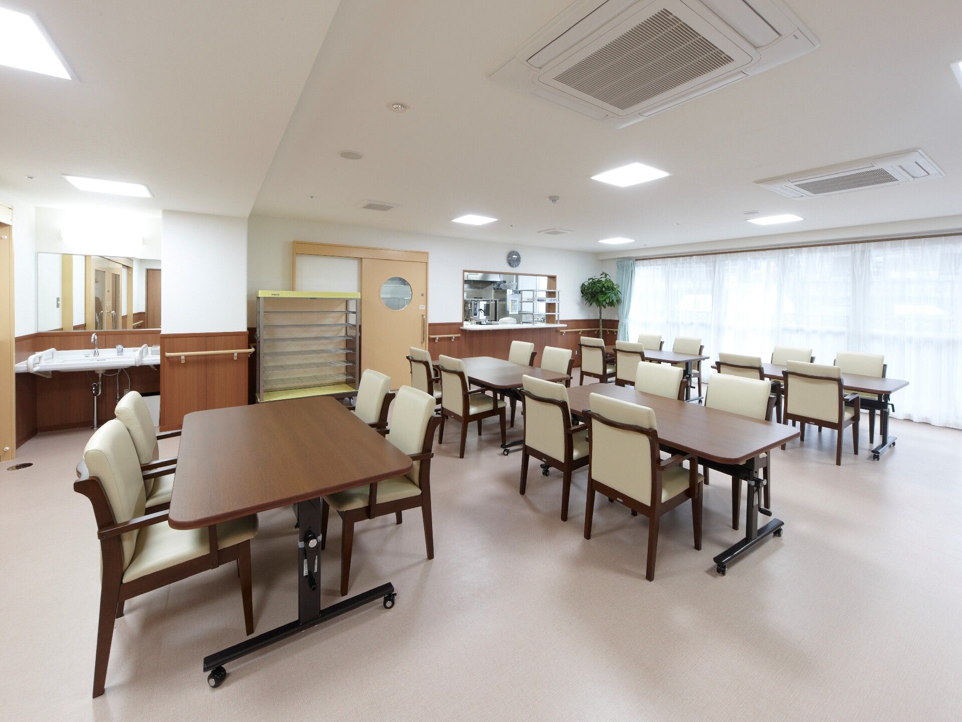 カーテンとカーテンが付いた大きな窓が設置された食堂にはデザインの異なるテーブルセットが複数置かれ、配膳者や観葉植物もある。