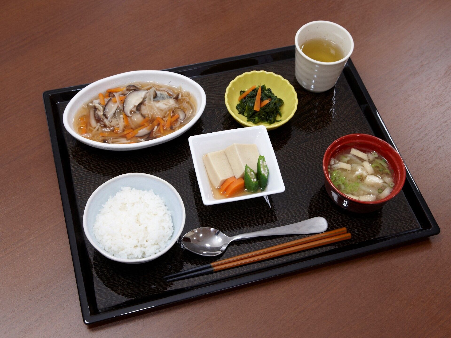キノコと人参のあんかけのようなものと青菜の和え物、みそ汁、高野豆腐の煮物、白いご飯などがトレーの上に並び、スプーンと箸が置かれいてる。