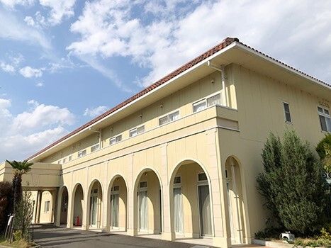 ベージュの外壁の2階建ての建物。日当たりのいい場所に建っていて、青空が広がっている。