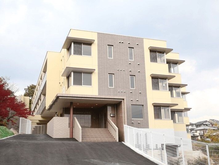 ベージュとブラウンの3階建ての建物。各フロアにベランダが付き、高台にあるため周りには高い建物はなく、紅葉した大きな木がある。