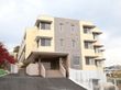 ベージュとブラウンの3階建ての建物。各フロアにベランダが付き、高台にあるため周りには高い建物はなく、紅葉した大きな木がある。