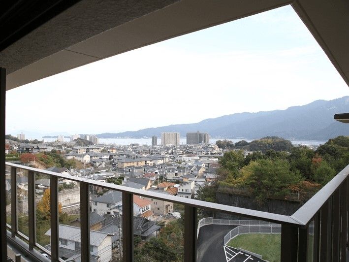 建物のベランダから周辺を眺めた風景。遠くには山々の連なる風景や海が見え、手前には住宅地が広がっていて、緑も残っている。