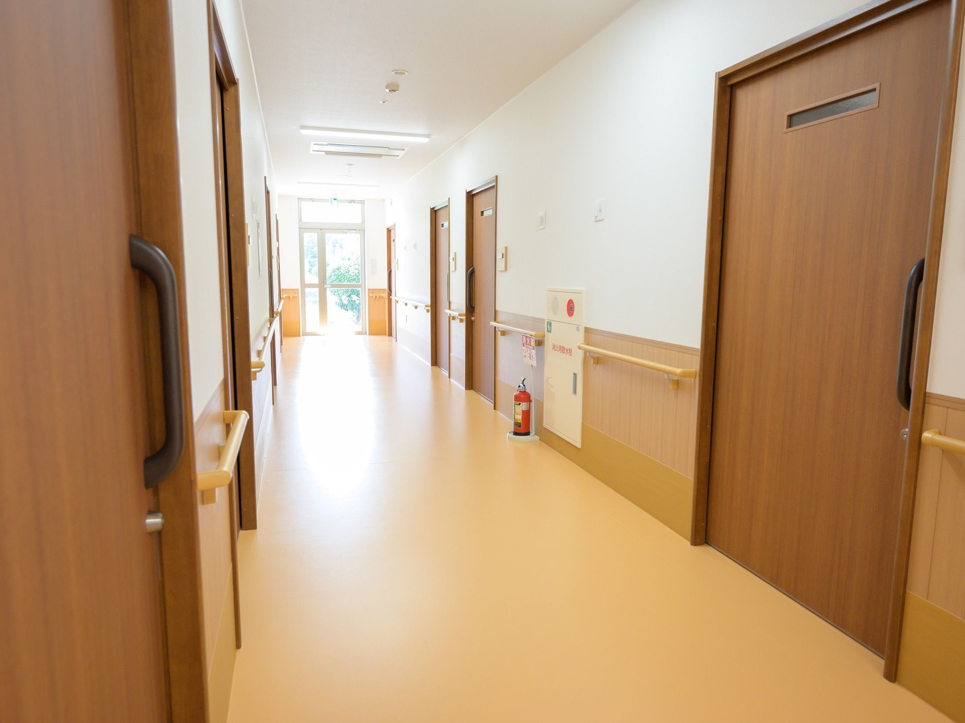 ゆとりのある幅の廊下があり、両側に居室の戸が並んでいる。木目調の引き戸で、壁には手すりが付いている。