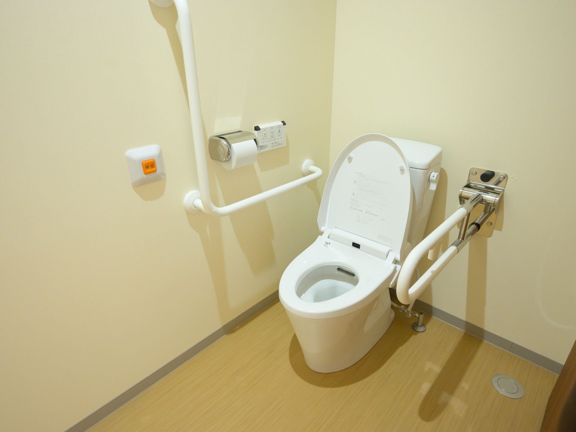 便座の両側に手すりが備え付けられたトイレがある。広々としていて、壁には緊急通報装置がある。