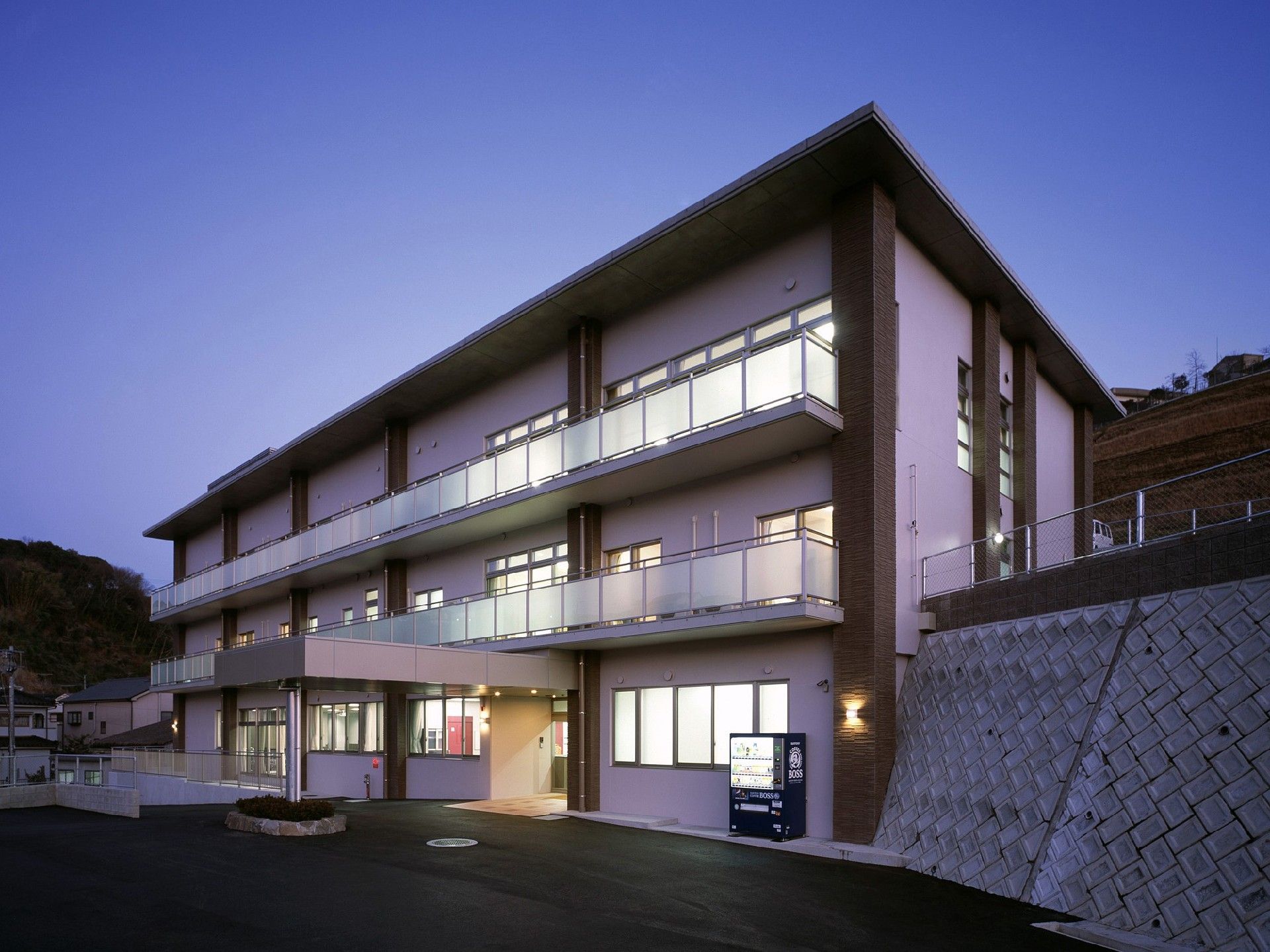 3階建ての建物の玄関にはスロープが付き、自動販売機もある。陽が沈み、あたりは薄暗い。