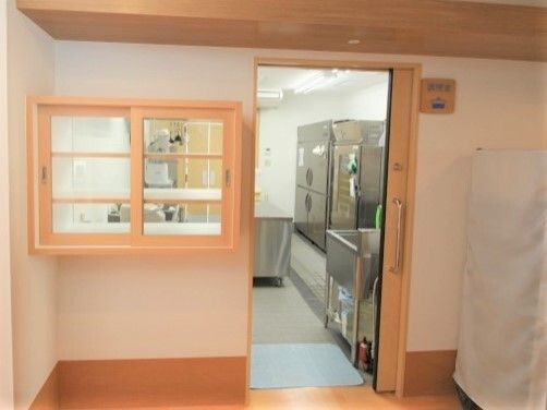 ステンレス製の調理器具が並んでいる広い厨房の出入り口にはスライド式のドアがあり、空いている。ドアの横にはトレーを並べることができる小窓もある。