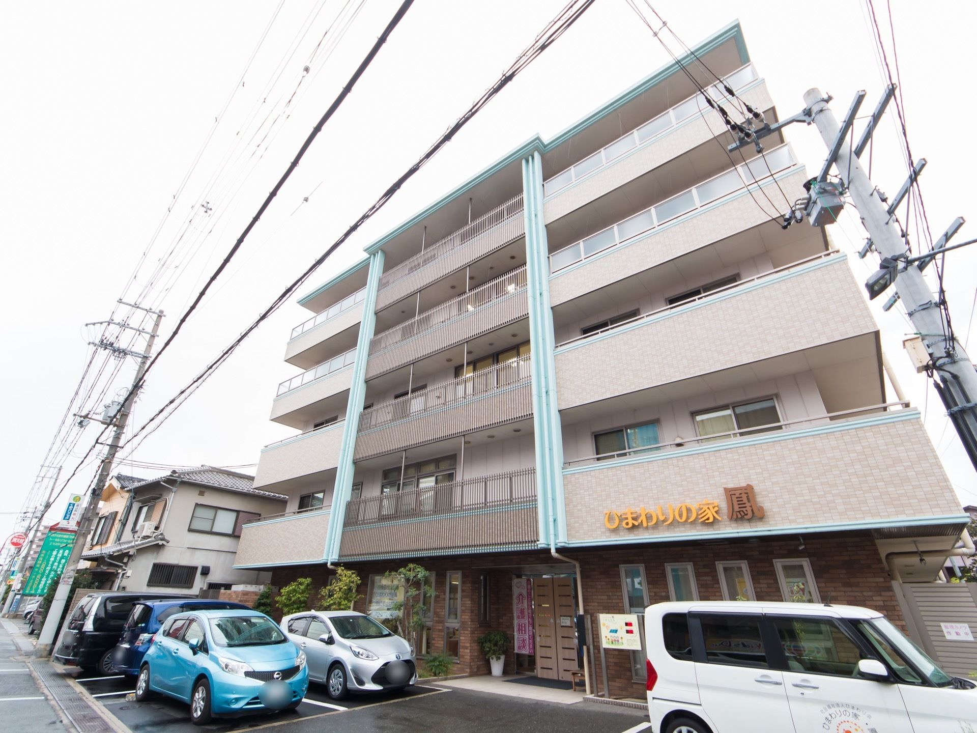 道路に面した敷地に建っている５階建ての施設である。建物の前に駐車スペースが確保されていて、車が何台か停まっている。