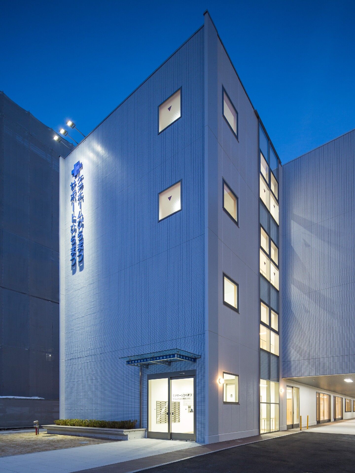 ライトアップされた4階建ての建物。玄関には自動ドアが付き、屋根が設置されていて、アプローチが続いている。
