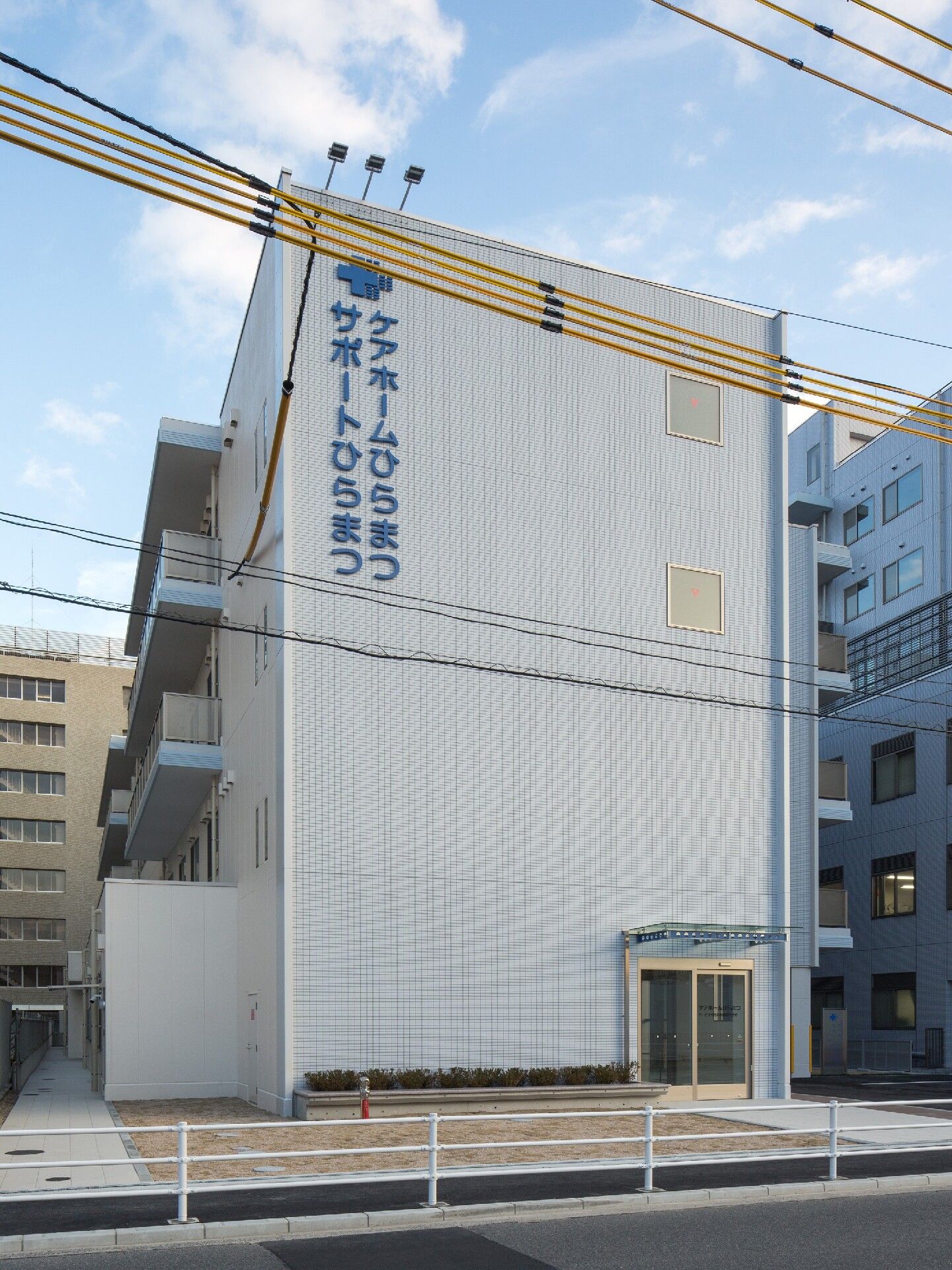 4階建ての建物は白を基調としていて、住宅名と併設事業所名が書かれている。周辺には背の高いビルもたくさんある。