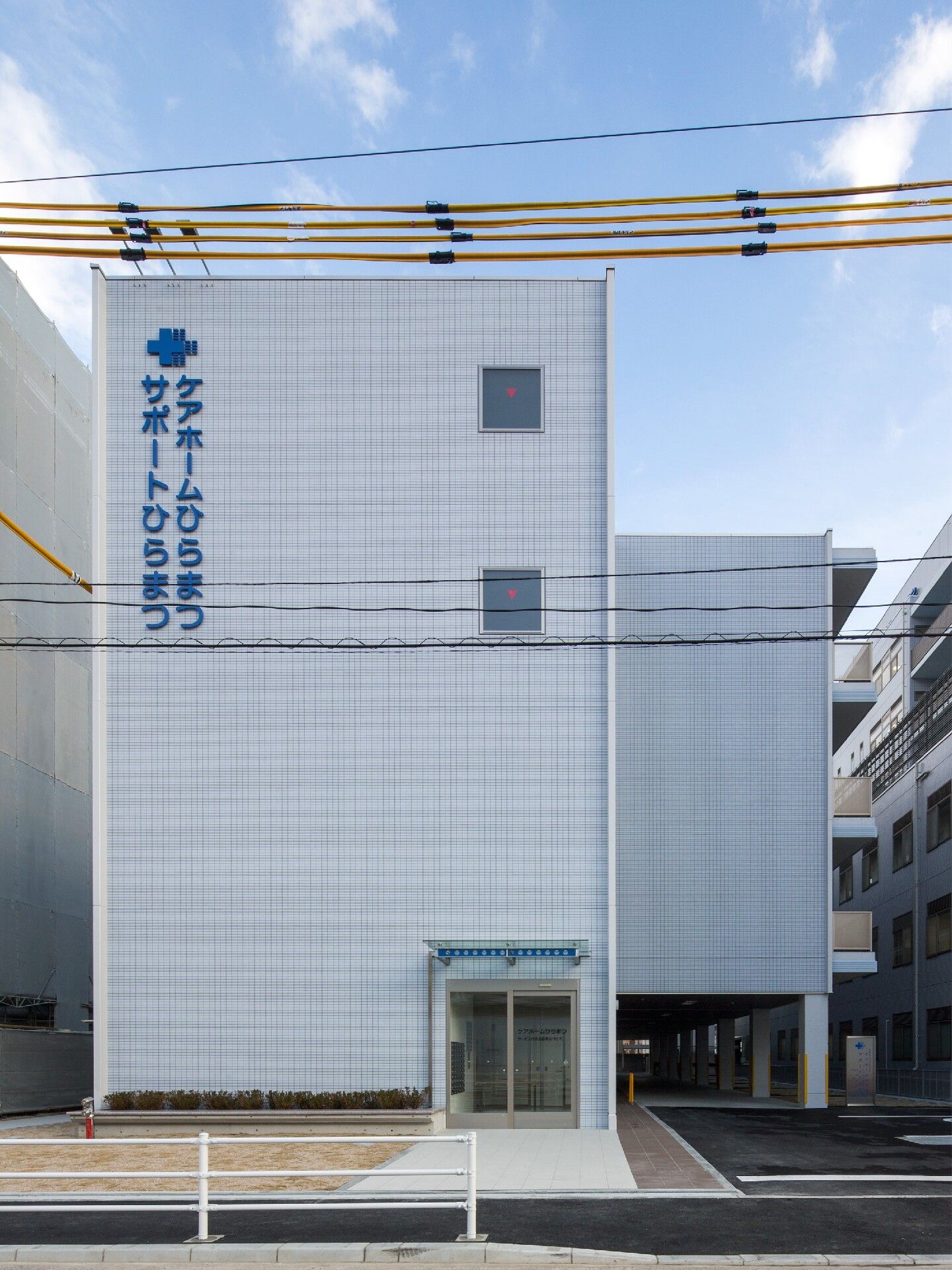 白い外壁には青い文字で住宅名と併設事業所名が書かれている。正面にある玄関には大きな屋根がある。