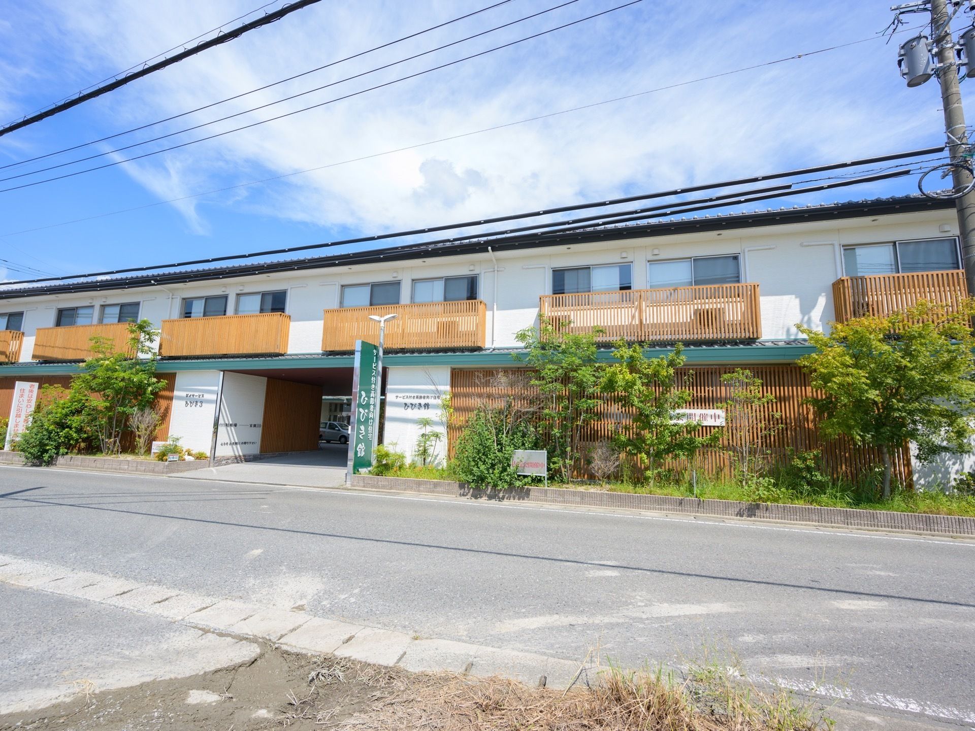 晴天の日の2階建ての施設の様子である。入口の両サイドに完備されている植え込みには、さまざまな種類の樹木が植わっており緑が鮮やかである。