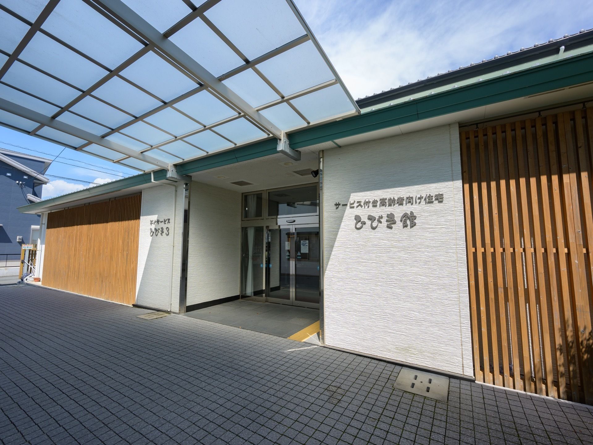 横幅広い施設建物の中央に完備された入口である。ガラスサッシの自動扉が設置され、上部には屋根がかかっている。
