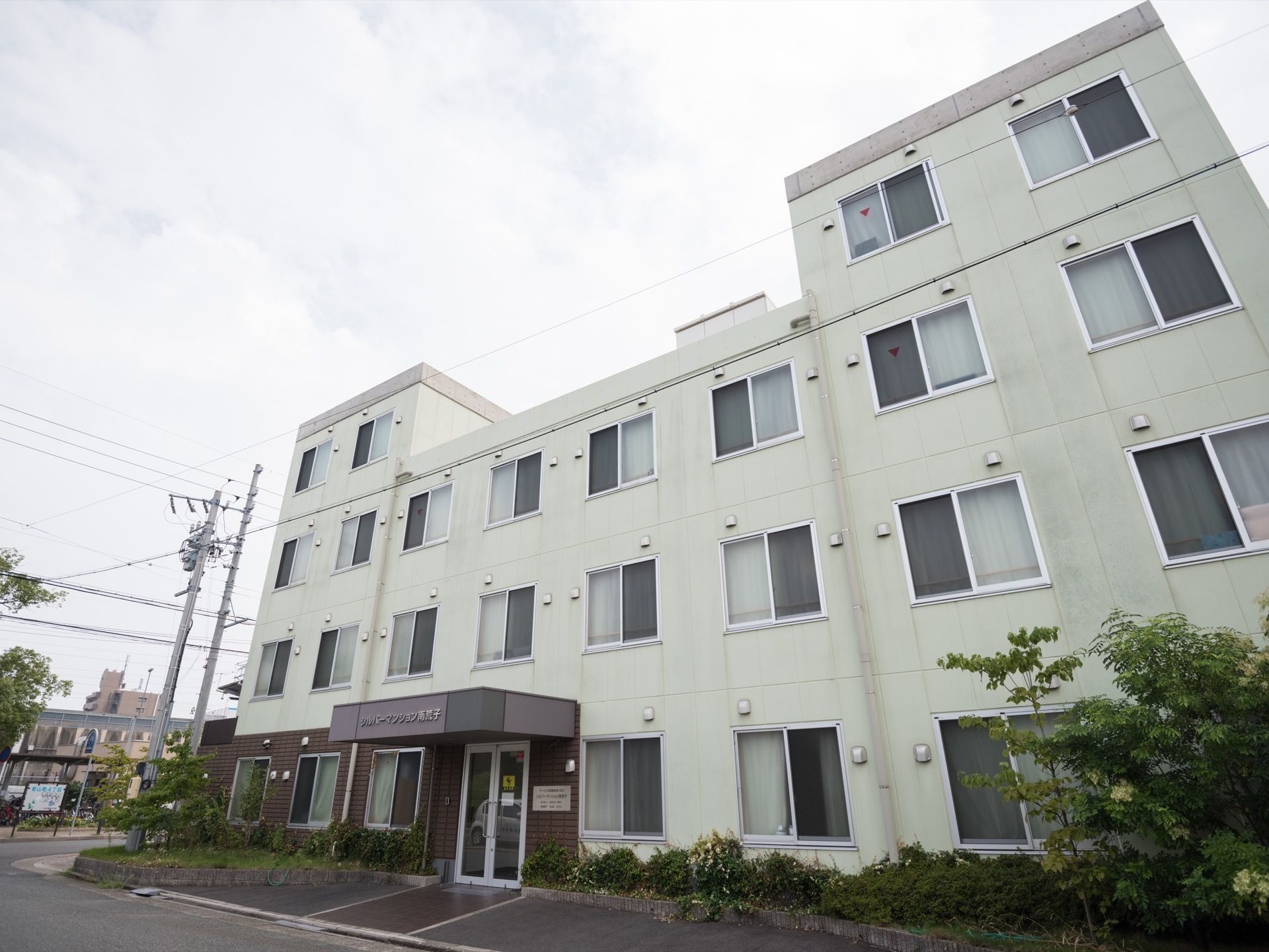 道路に面した場所に、白と茶色の４階建て建造物がある。駐車場が施設の目の前にあり、まわりに草木が植えられている。