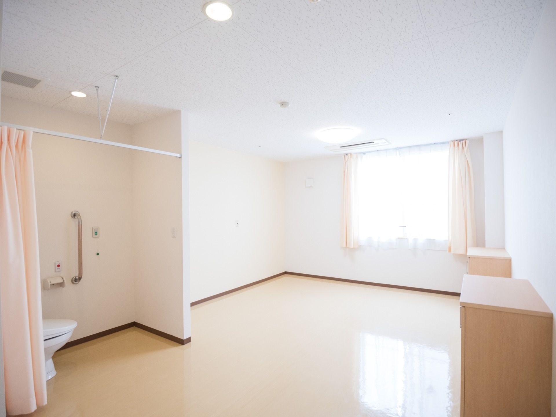 窓が付いた部屋の一角がカーテンで仕切られており、蓋の付いた洋式便器が据えてある。壁面にステンレス製の手すりとナースコールが設置されている。