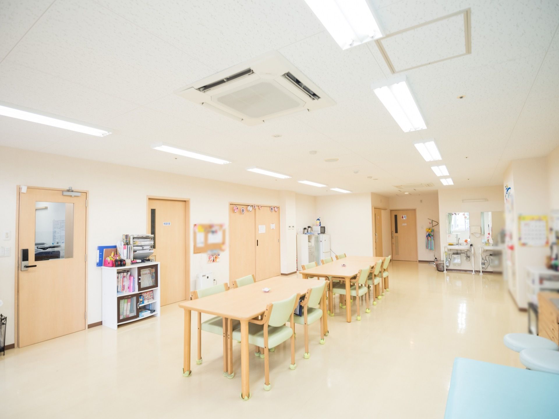 広いスペースに、木製テーブルや介護用椅子が並べられている。右奥に洗面スペースが設置されているのが見える。