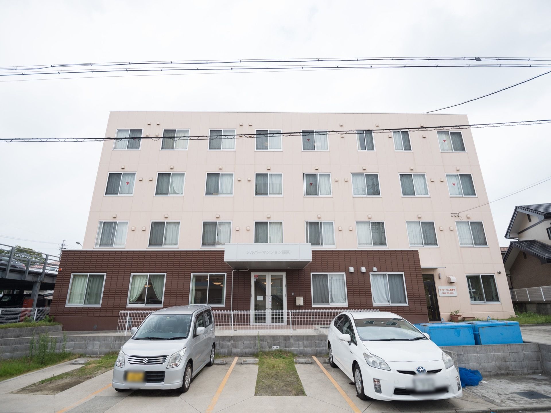 窓が並んだ4階建ての施設が建っている。手前に区画された駐車場があり、建物の左側に立体駐車場が見える。