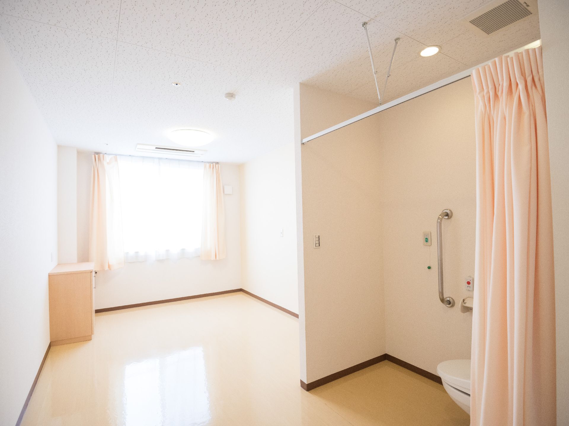 カーテンが間仕切りになるトイレが付いている、白い内装で明るい居室