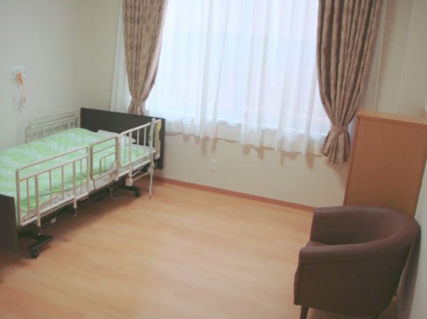 レースカーテンとカーテンが付いている大きな窓があるフローリングの部屋には、寝具付きのベッドがあり、ベッドサイドには緊急通報装置が付いている。