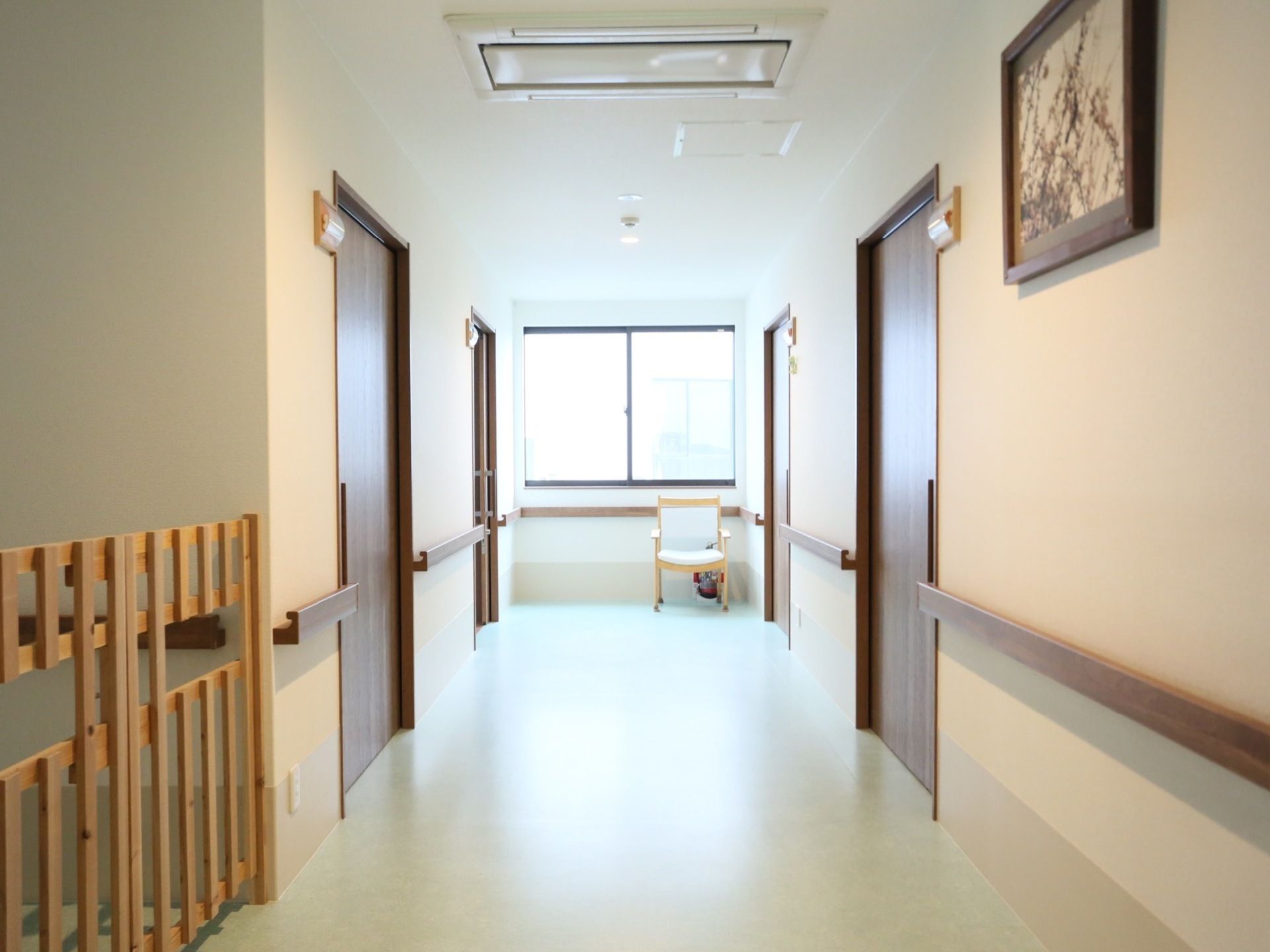 廊下の両側の壁に、板状の手すりが設置されている。つきあたりの窓の前に、背もたれとひじ掛けがついた椅子が１つ置かれている。