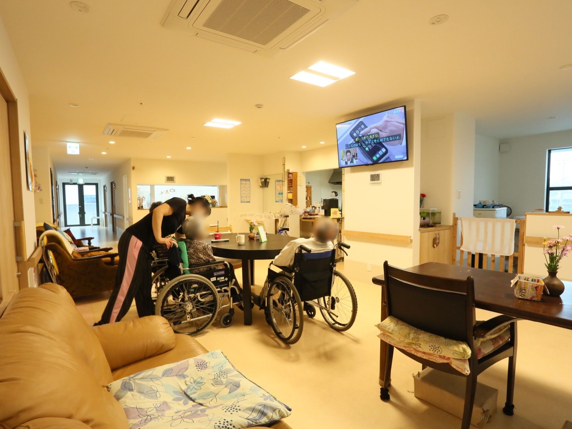 車椅子をご利用の入居者様が２名、共有スペースのテーブルについている。女性スタッフが入居者様のお世話をしている。
