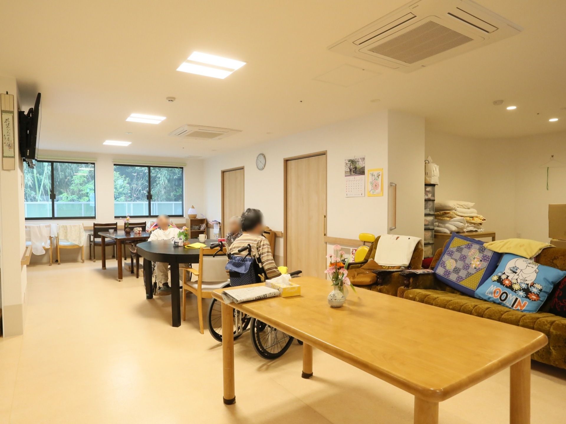 共有スペースには、いろいろな形のテーブルが置かれている。入居者様が３名集まっている。窓からは木々が見える。