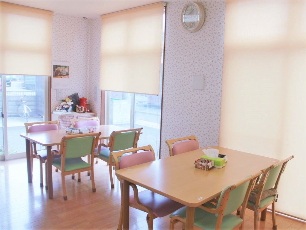 緑とピンクの椅子が2脚ずつセットされたダイニングテーブルが複数あり、ぬいぐるみやカレンダーも飾られている。