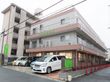 道路側に駐車スペースがあり、車が数台停められている。その奥に建物が伸びており、建物脇に玄関への通路が設けられている。