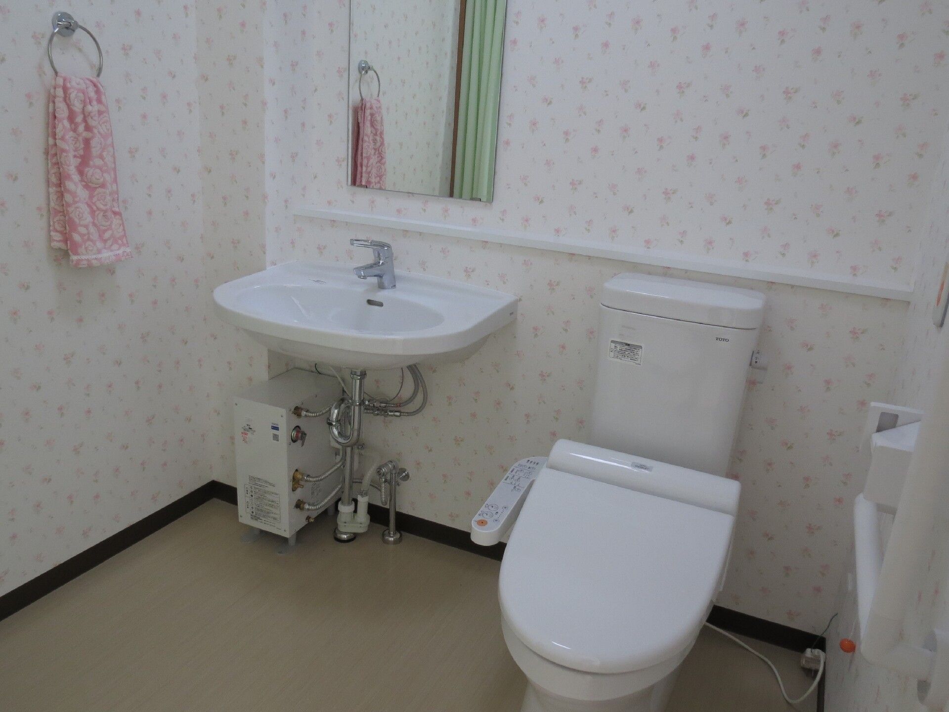 広いトイレには車いす対応の洗面所があり、壁にはオレンジの呼び出しボタンやタオル掛け、手すりなども付いている。