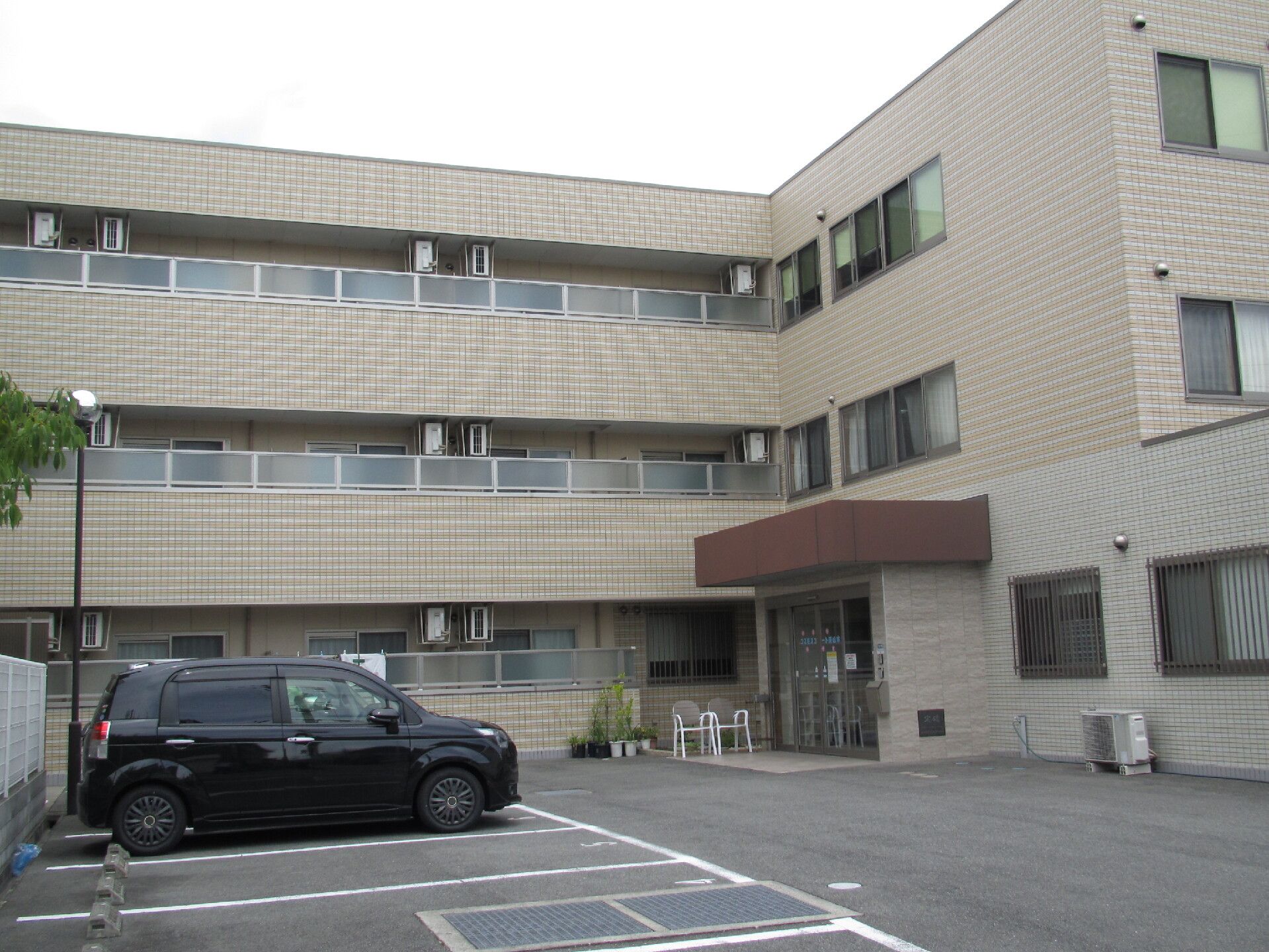 各フロアにベランダがあり、鉄骨造りの建物はベージュ系でまとめられた3階建て。広い駐車場があり、数台の車が止められている。