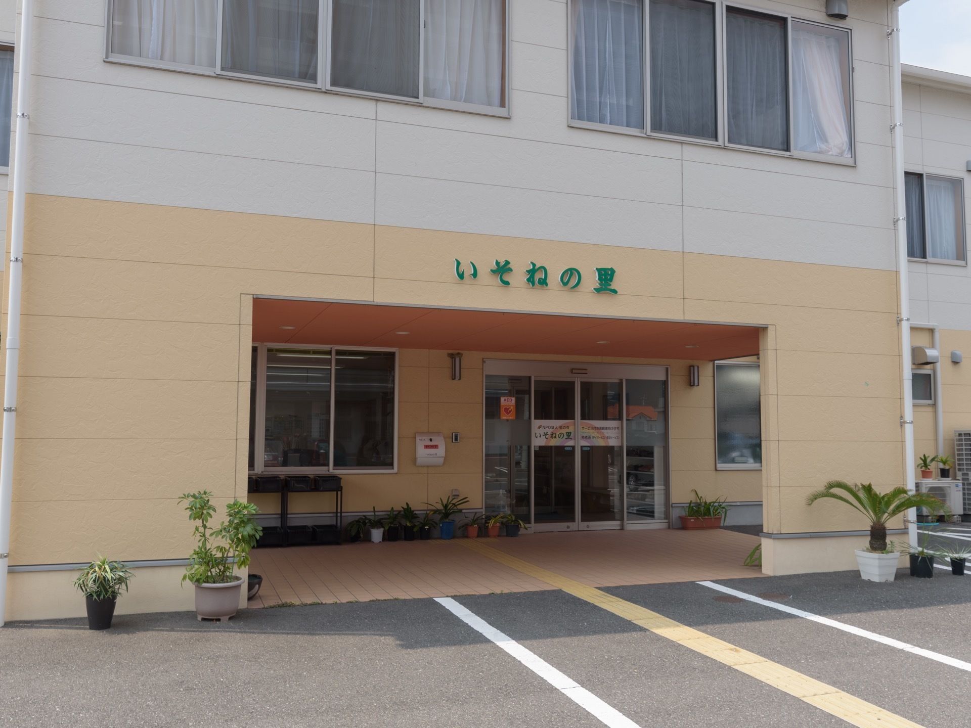 自動ドア横に、複数個の鉢植えやプランターが並べられている。玄関上の外壁には、緑色で施設名が書かれている。