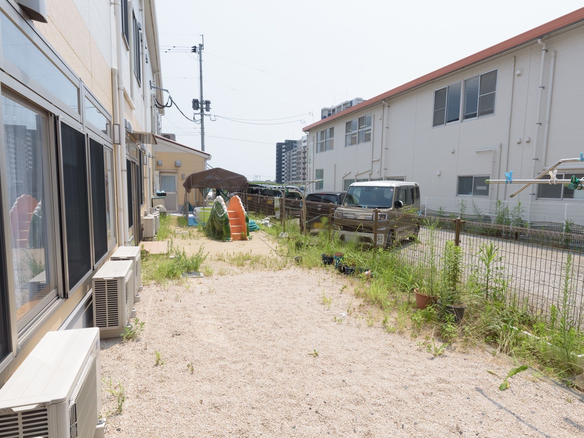 ２階建てのグループホームで、外壁沿いにエアコンの室外機が複数台配置されている。遠くに、背の高いマンションが建っているのが見える。
