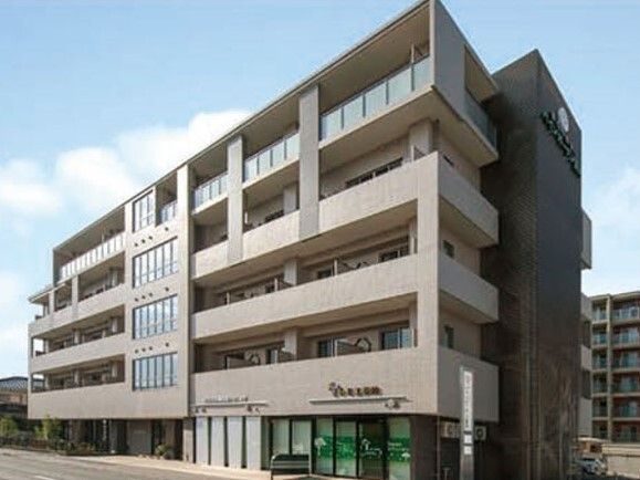 グレーの5階建ての建物はベランダが付き、重厚感がある。周辺にはマンションのような背の高い建物もある。