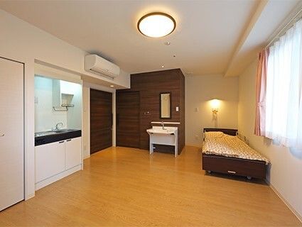 キッチンが付いたフローリングの部屋にはベッドがあり、ベッドサイドには緊急通報装置がある。