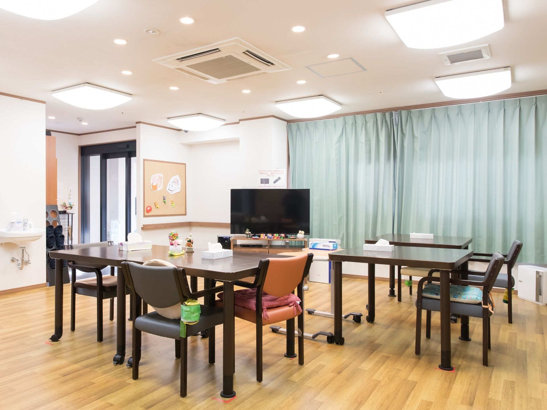 天井に複数の照明が設置された明るい食堂である。テレビやテーブル、椅子が配置されている。左奥に玄関がある。