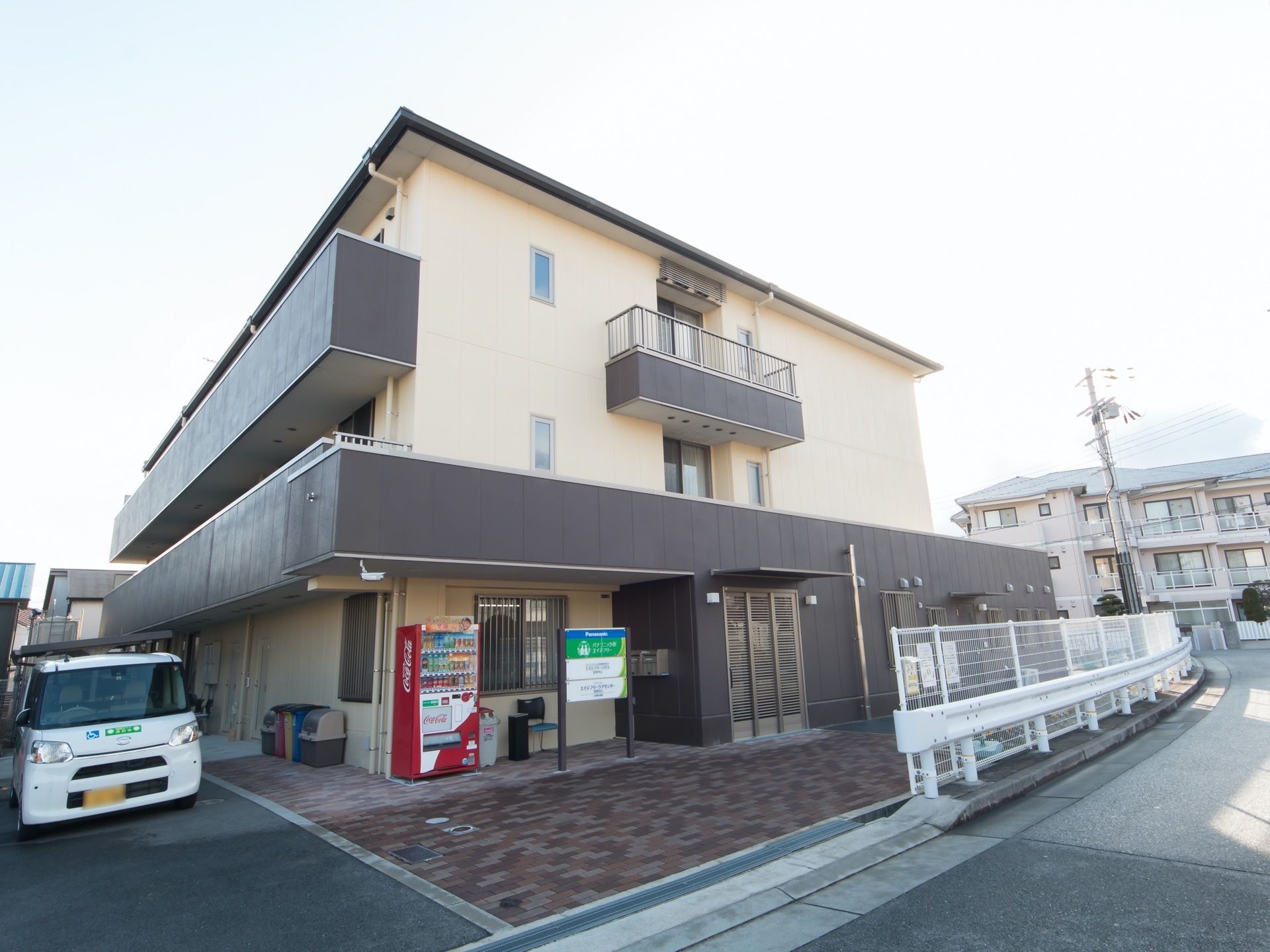 ３階建ての施設建物である。玄関の脇には、施設名や説明が書かれた看板や、自動販売機が設置されている。施設と車道の間にはゆとりがある。