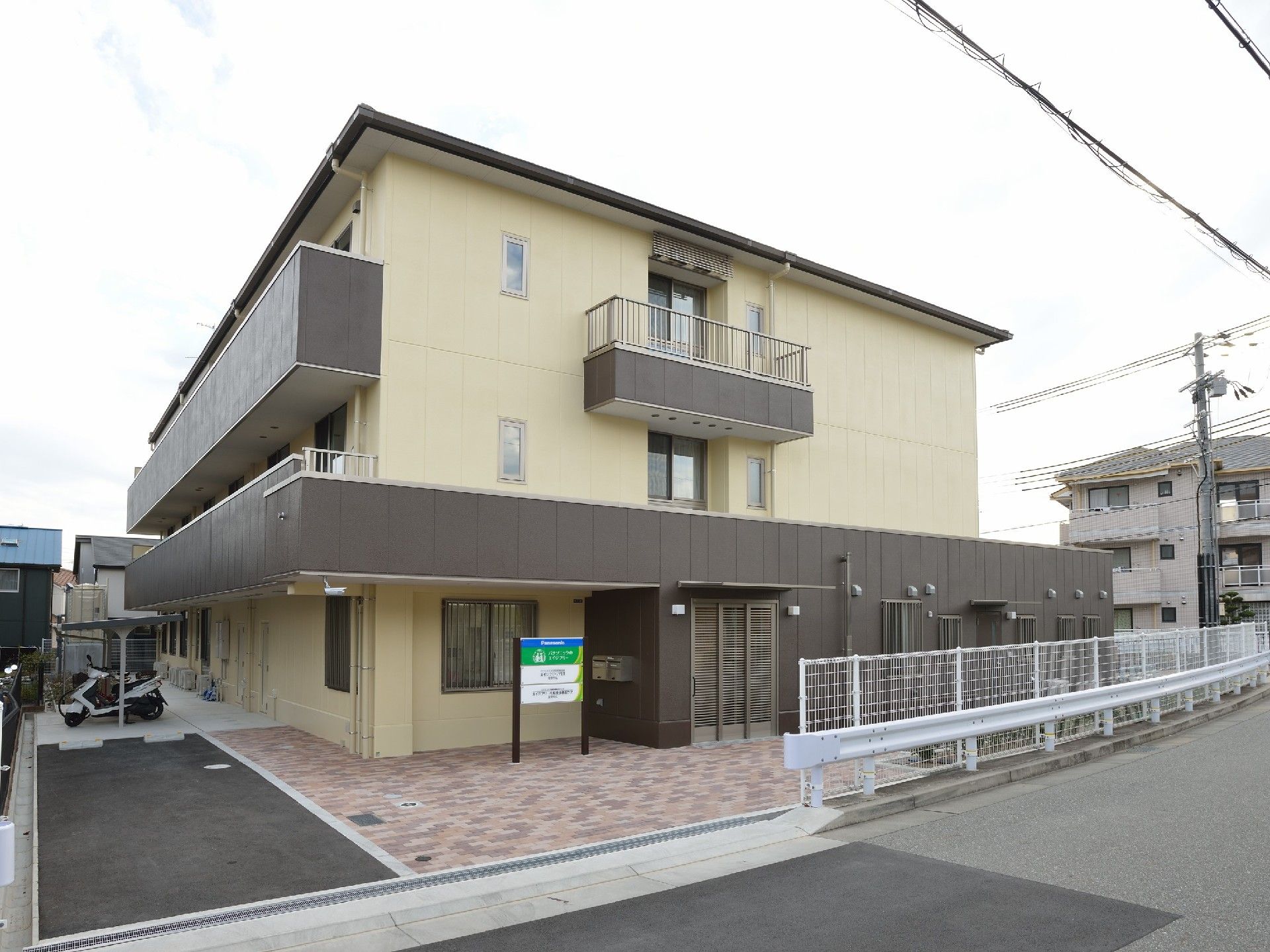 駐輪場がついた敷地内には3階建ての建物があり、玄関の近くに看板も設置されている。