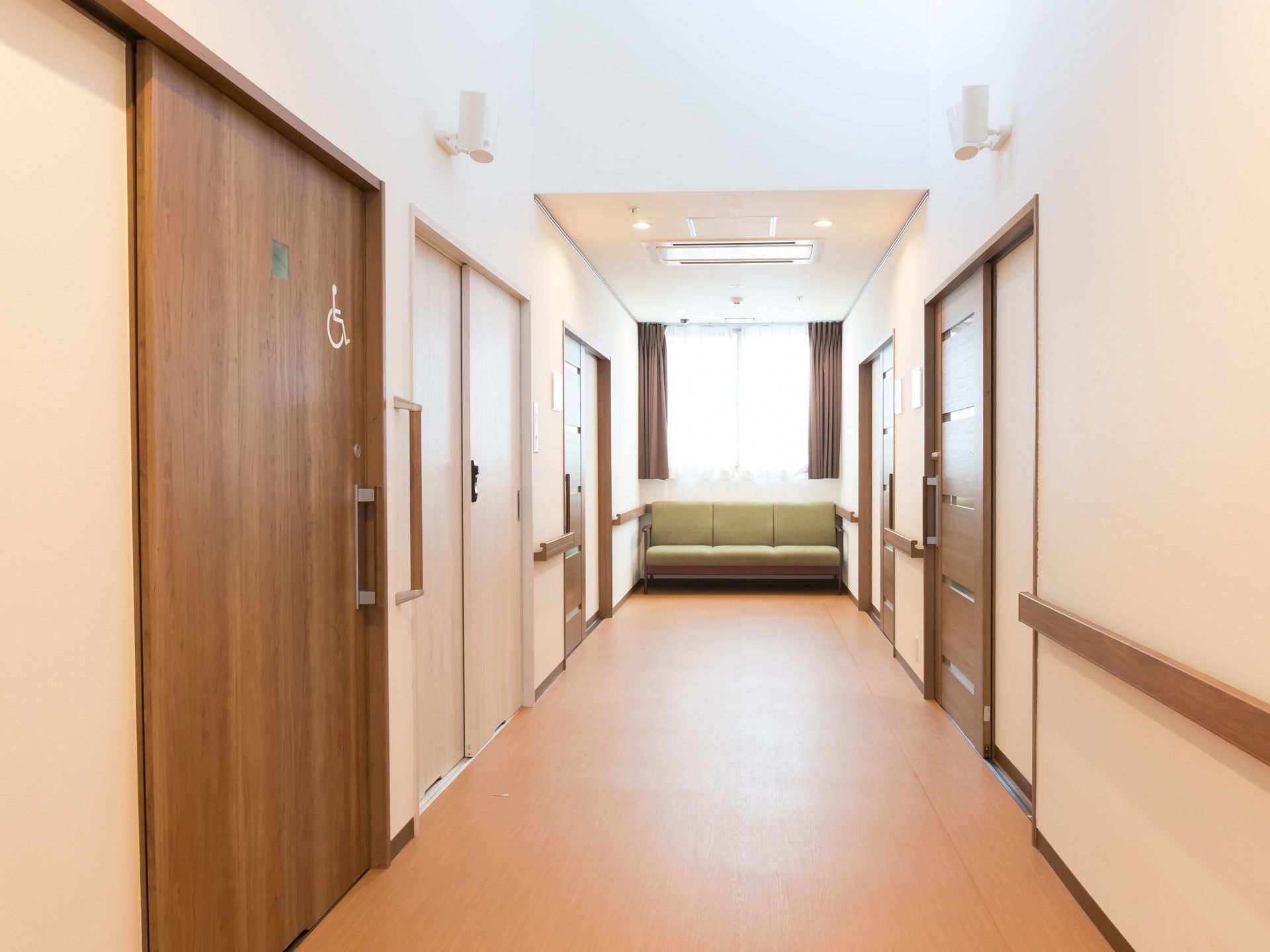 木目調の手すりが備え付けられた廊下が続いている。突き当たりには窓があり、その下には緑色のソファが置かれている。